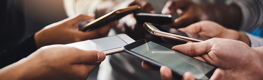 Close-up of several hands of different skin tones holding smartphones in a circle, screens facing inward, suggesting social interaction, sharing, or collaboration. The background is blurred, focusing attention on the phones and hands.