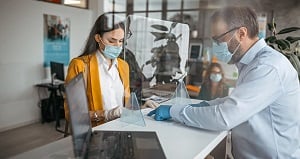 A woman and a man, both wearing face masks, stand on opposite sides of a counter separated by a clear plastic barrier. The man, wearing gloves, points to a document while the woman listens. A laptop is open on the counter in a bright office setting.