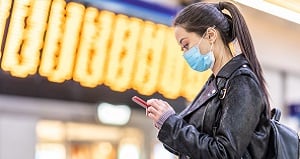 A young woman wearing a blue surgical mask and a black leather jacket stands indoors, looking at her smartphone. Blurred, bright yellow electronic signage is visible in the background, suggesting a public transit or train station setting.