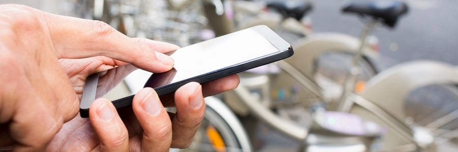 A close-up of a person’s hands using a smartphone outdoors, with several blurred bicycles in the background, suggesting a possible urban bike-sharing setting. The focus is on the phone and hands, with the background out of focus.