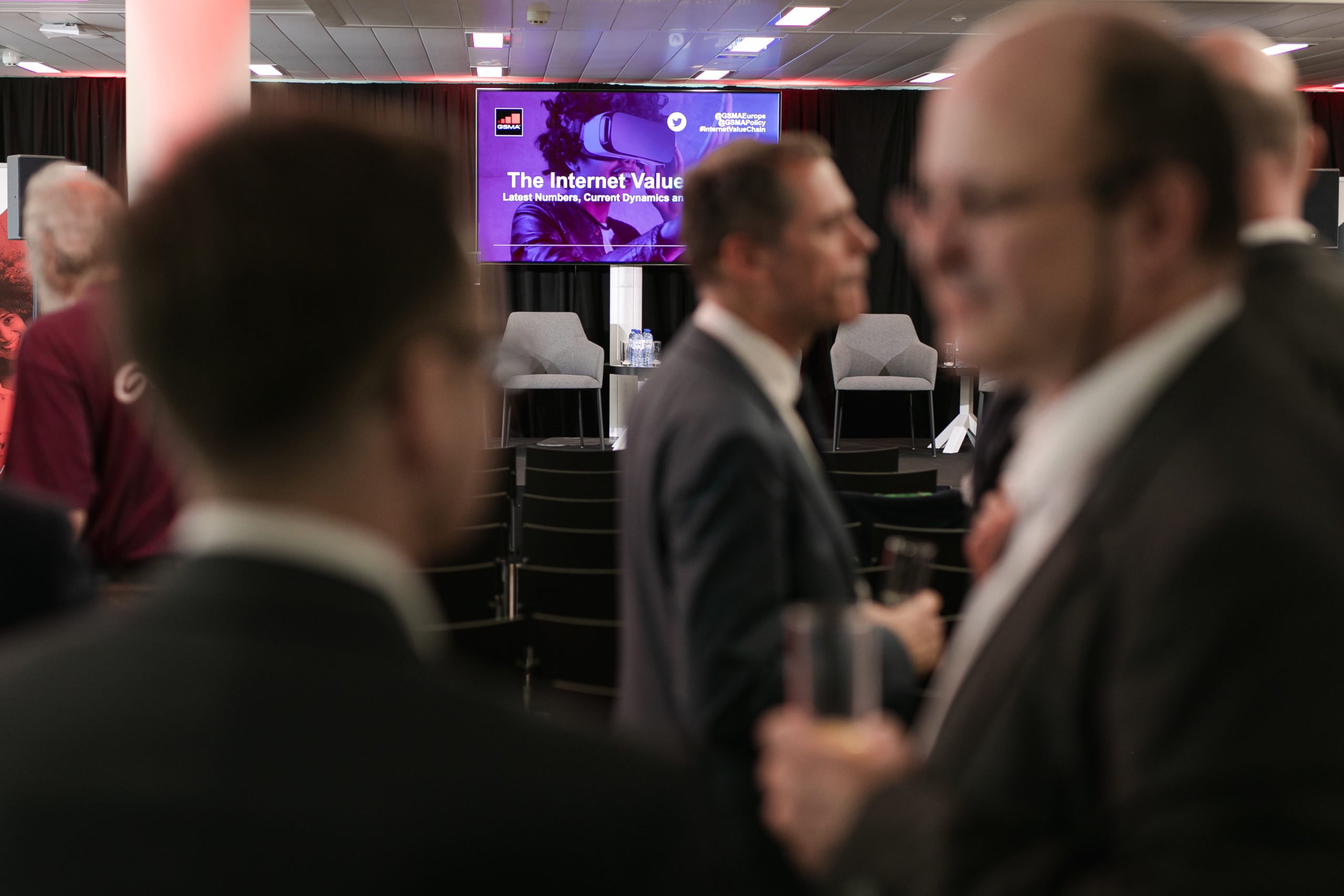 A blurry foreground shows people in suits conversing at an event. In the background, a clear screen displays “The Internet Value,” suggesting a conference or seminar setting. Empty chairs and a stage are visible behind the attendees.