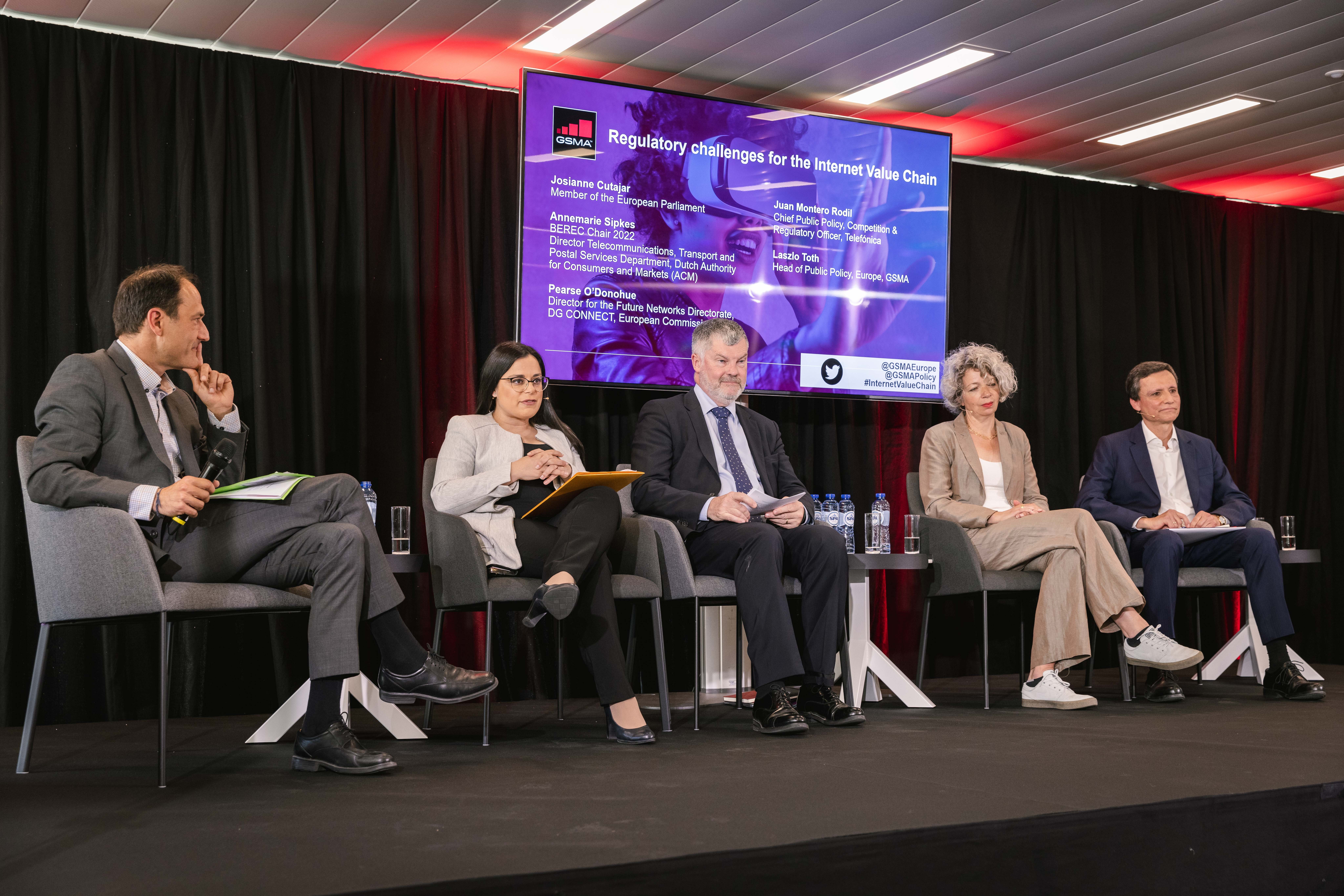 Five professionals sit on stage in armchairs, engaged in a panel discussion. Four men and one woman face an audience. Behind them, a large screen displays the event topic: "Regulatory challenges for the Internet Value Chain," with names and titles of speakers.