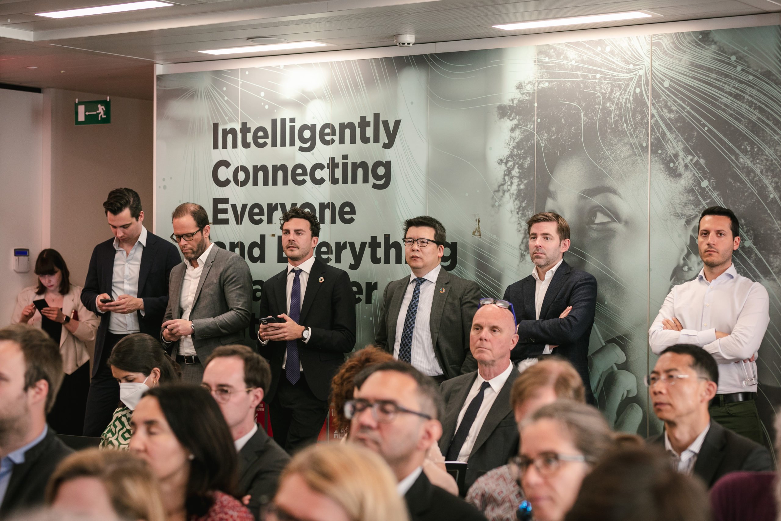 A group of professionally dressed people stand against a wall at a business event. The wall features a large graphic with the words “Intelligently Connecting Everyone and Everything.” Many seated attendees face forward in the foreground.