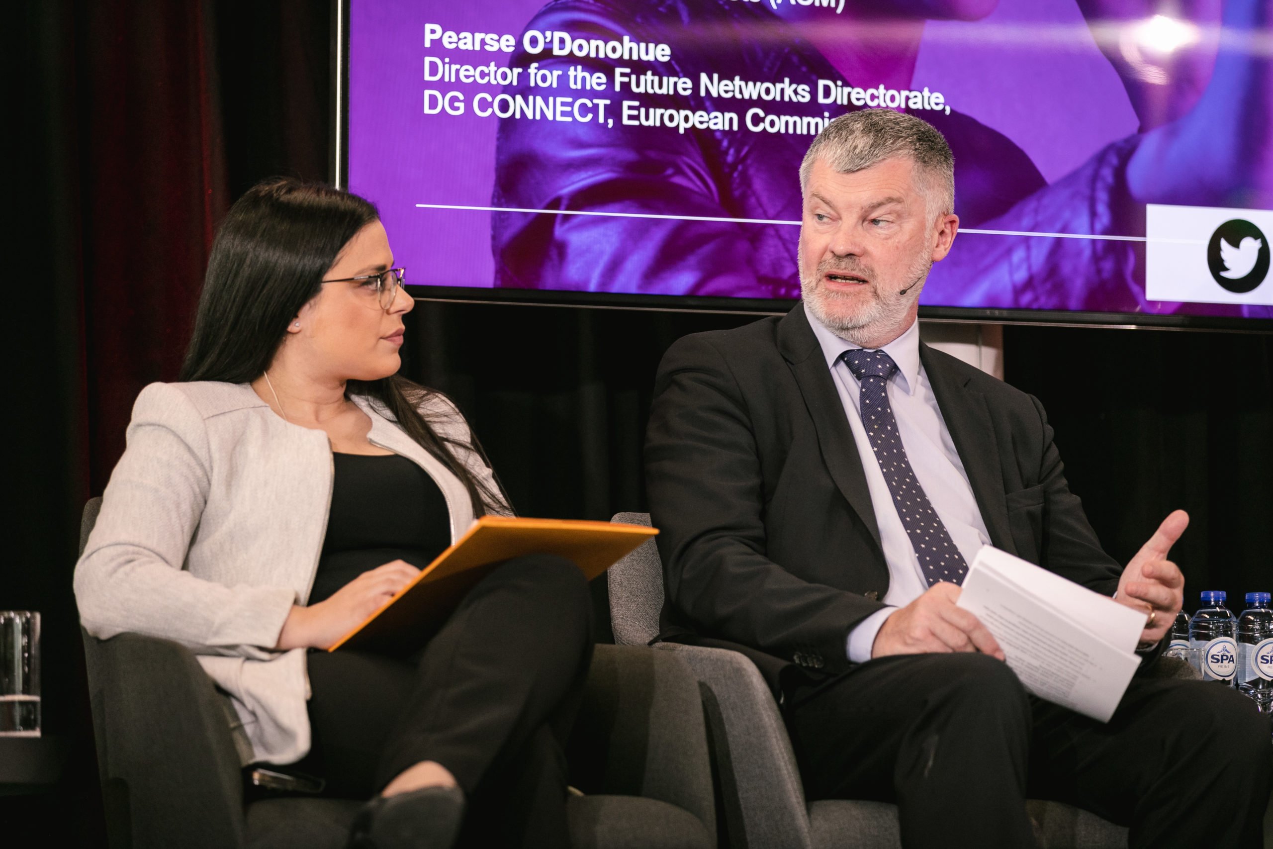 A woman in a light blazer and glasses holds an orange folder while listening to a bearded man in a suit speaking. They are seated onstage at an event, with a purple screen in the background highlighting Pearse O’Donohue’s name and title.
