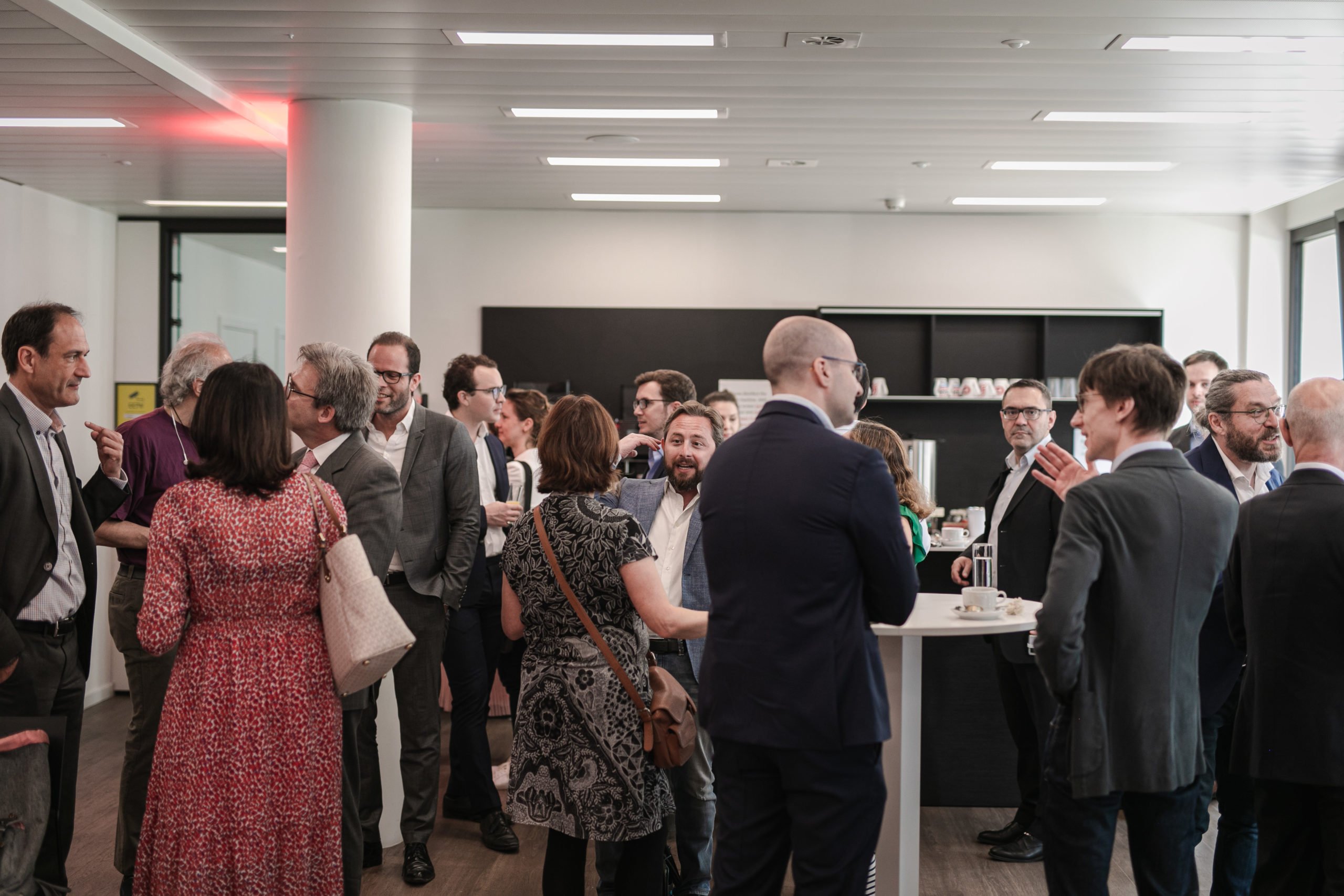 A group of professionally dressed people mingle and converse in a modern office space. Some hold drinks or coffee cups. The room is bright with minimal decor, featuring large windows, white walls, wooden floors, and a black shelving unit in the background.