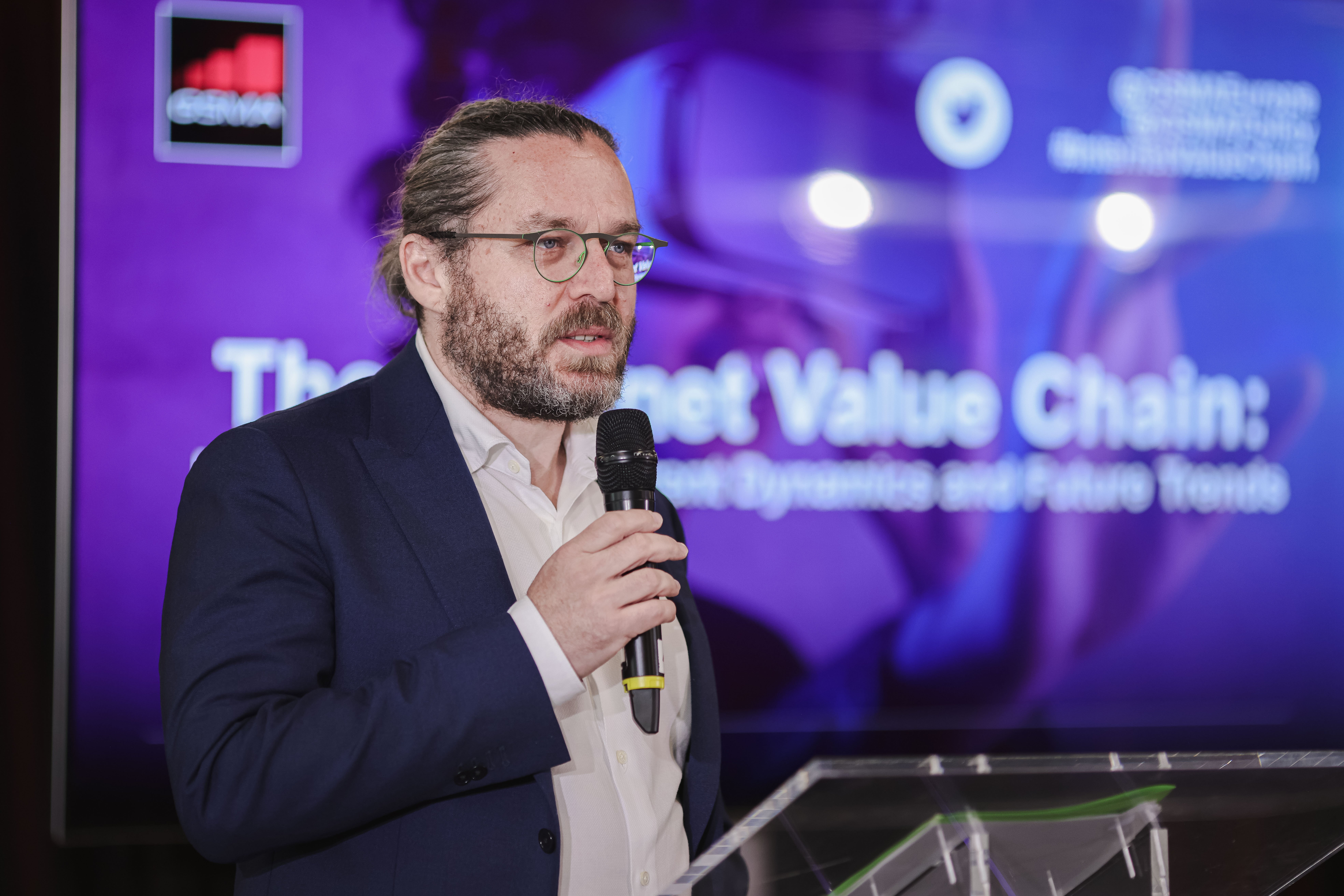 A man with glasses, a beard, and long hair tied back, wearing a navy suit and white shirt, speaks into a microphone at a clear podium. Behind him is a purple screen displaying text and logos related to the Internet Value Chain.
