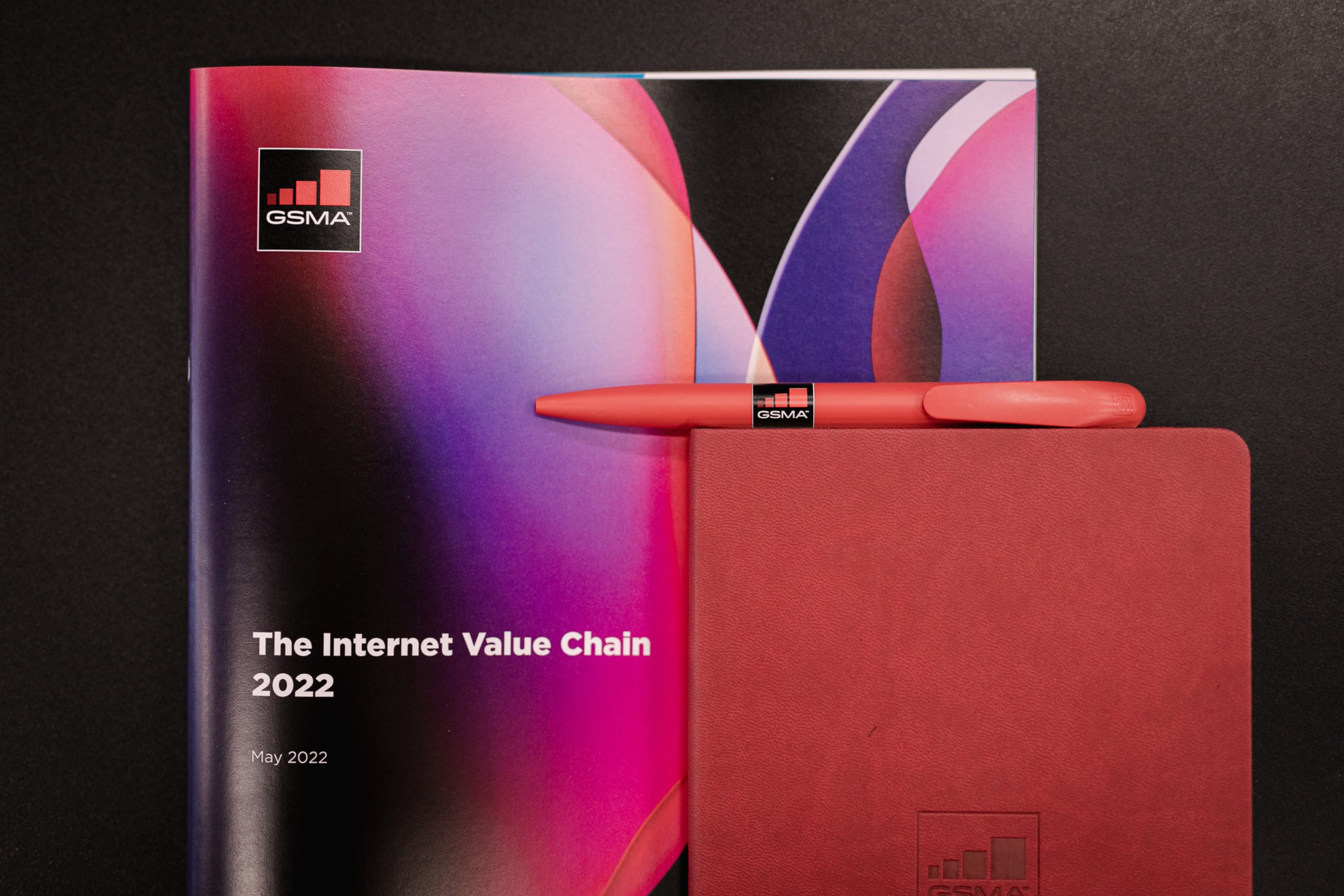 A GSMA-branded booklet titled "The Internet Value Chain 2022" lies on a black surface. A red GSMA pen and a red notebook with the GSMA logo are placed on top, with vibrant abstract graphics featured on the booklet’s cover.