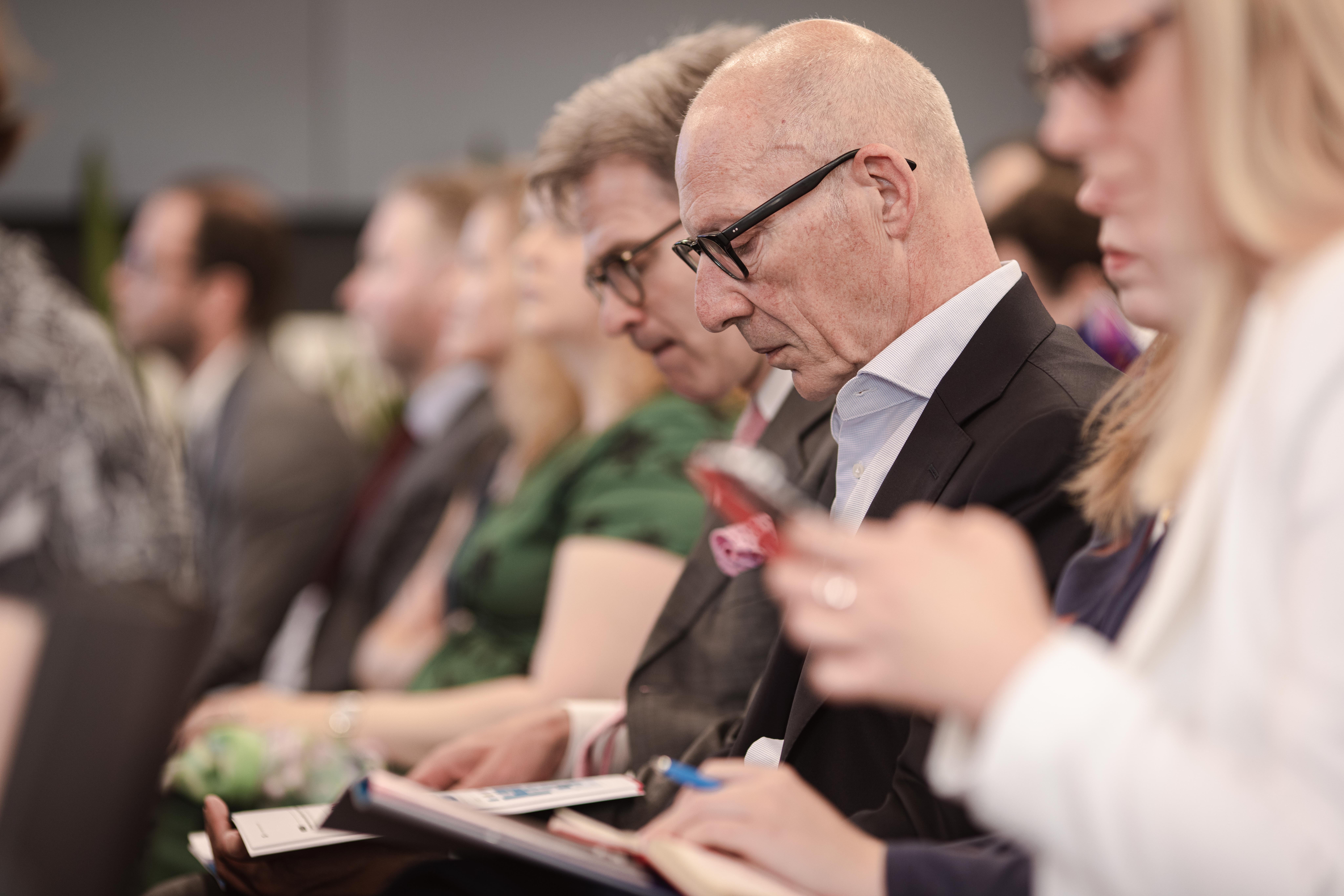 A group of people sit closely together in a conference or seminar setting. Most are dressed in business attire and appear focused, with some taking notes or looking at their phones and notebooks. The atmosphere is serious and attentive.