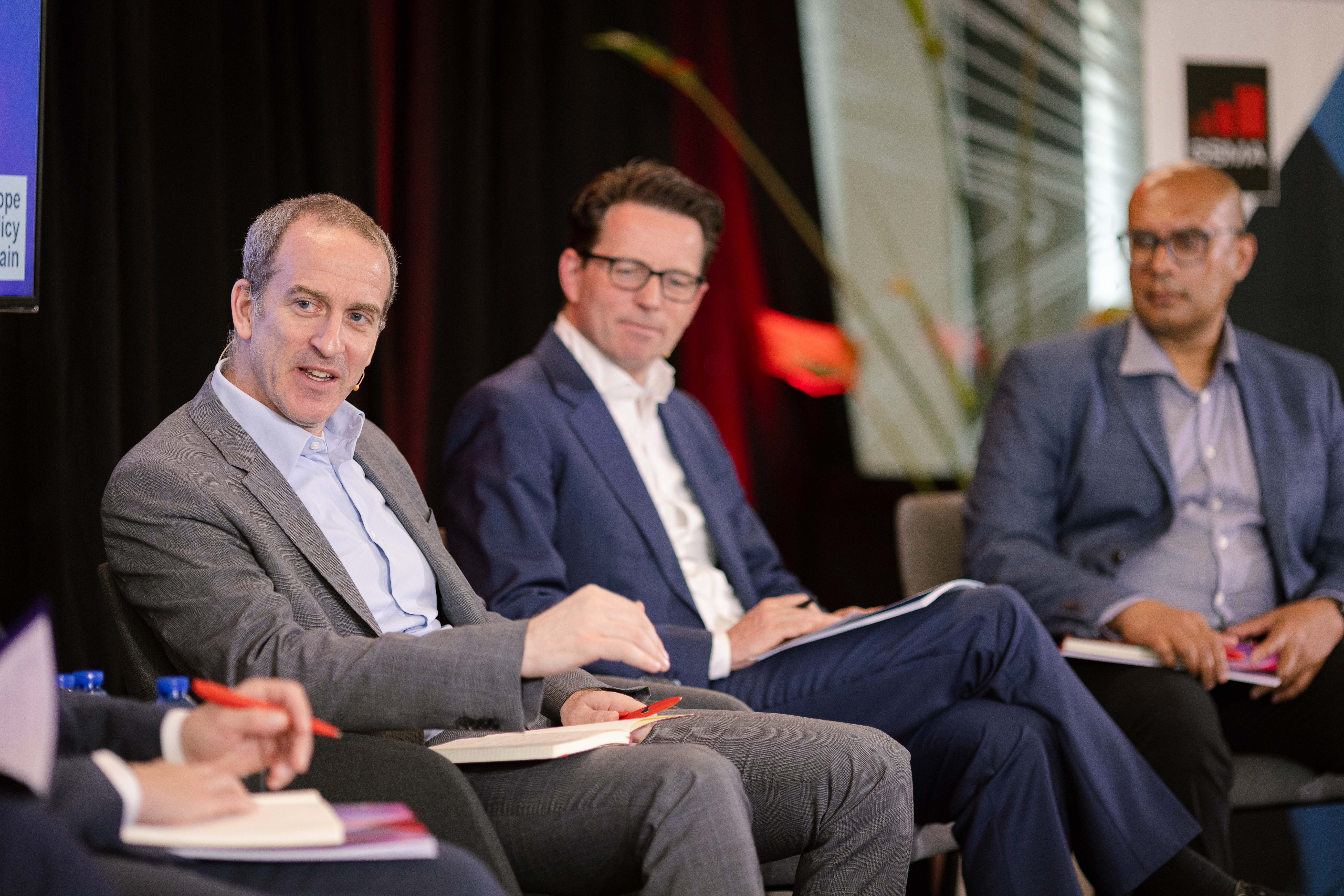 Three men sit on a panel in business suits, engaged in discussion. One man gestures while speaking, with the others listening attentively. Notebooks, pens, and papers are visible in their laps. The background features dark curtains and modern office decor.