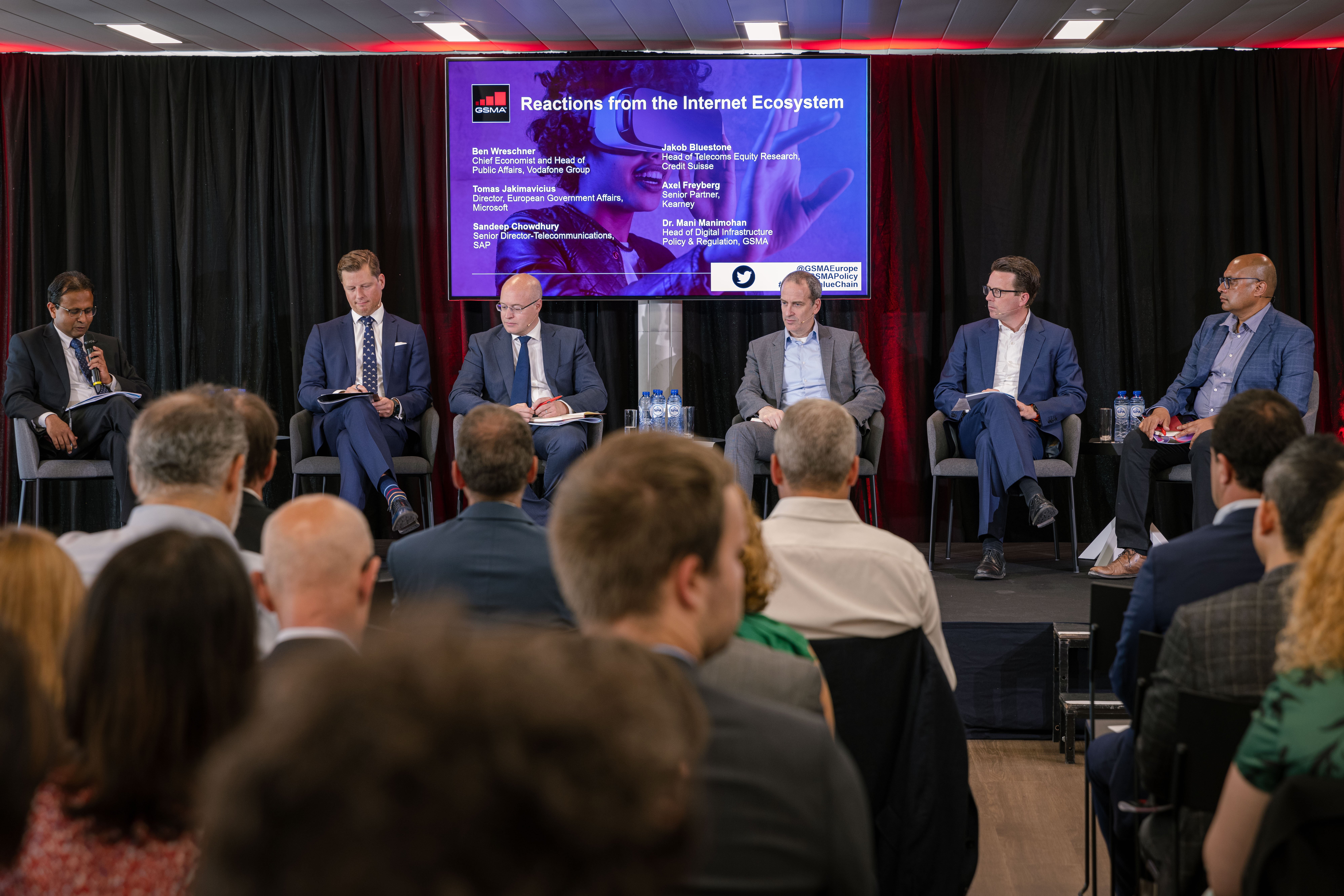 Six men in business attire sit on a stage panel, holding microphones or notes, with an audience in front. Behind them, a large screen displays the event title "Reactions from the Internet Ecosystem" alongside participant names and event details.
