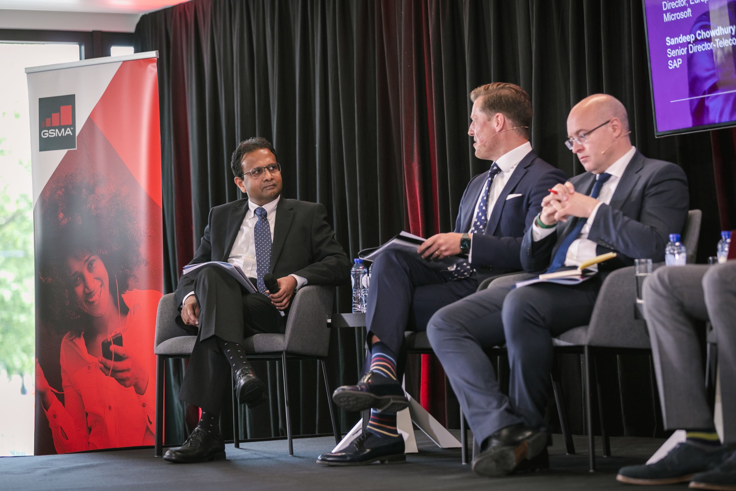 Four men in business attire sit on a stage for a panel discussion. One man on the left looks at the other three, who hold papers. There’s a GSMA banner with a woman’s photo on the left and a black curtain and purple screen in the background.