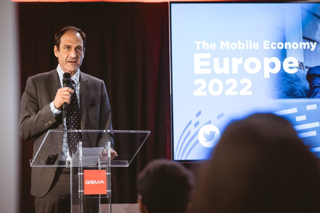 A man in a suit speaks into a microphone at a clear podium with a red GSMA logo. Behind him, a screen displays "The Mobile Economy Europe 2022" in bold white letters on a blue background. Audience members are visible in the foreground.