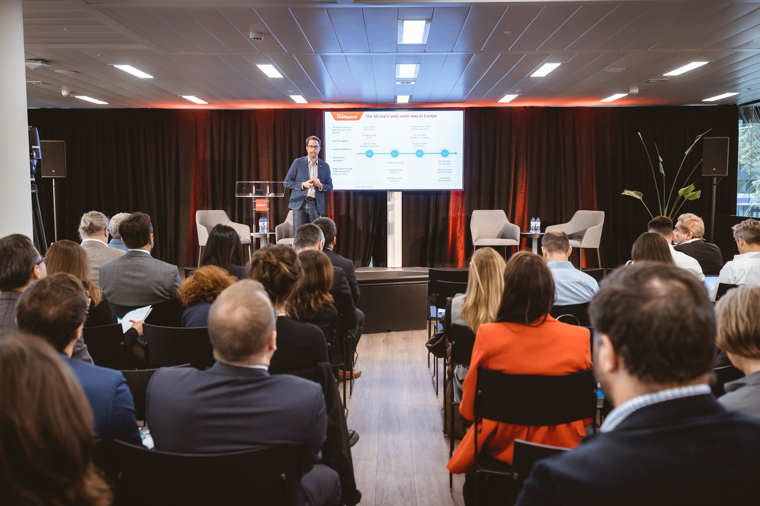 A man in a suit stands on stage giving a presentation to a seated audience. Behind him is a screen displaying a slide with graphs and text. The stage has two empty chairs, a small table, and is framed by dark curtains with orange lighting.