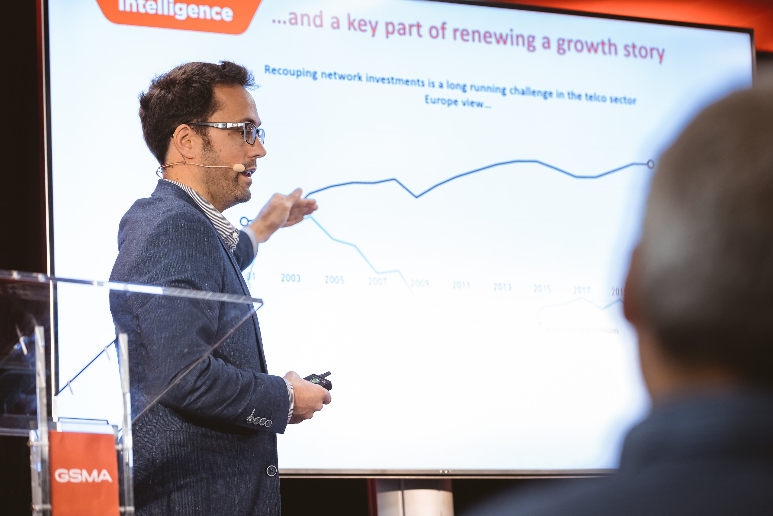 A man in a blue suit and headset stands at a podium, pointing to a projected line graph during a presentation. The slide’s visible title reads, “…and a key part of renewing a growth story.” The setting appears to be a professional conference.
