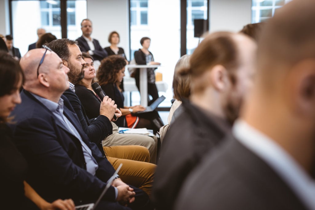 Audience members sit in rows at a conference or seminar. One man in the foreground holds a microphone, poised to speak. People appear engaged, listening attentively. Blurred figures and large windows are visible in the background, adding a professional setting.