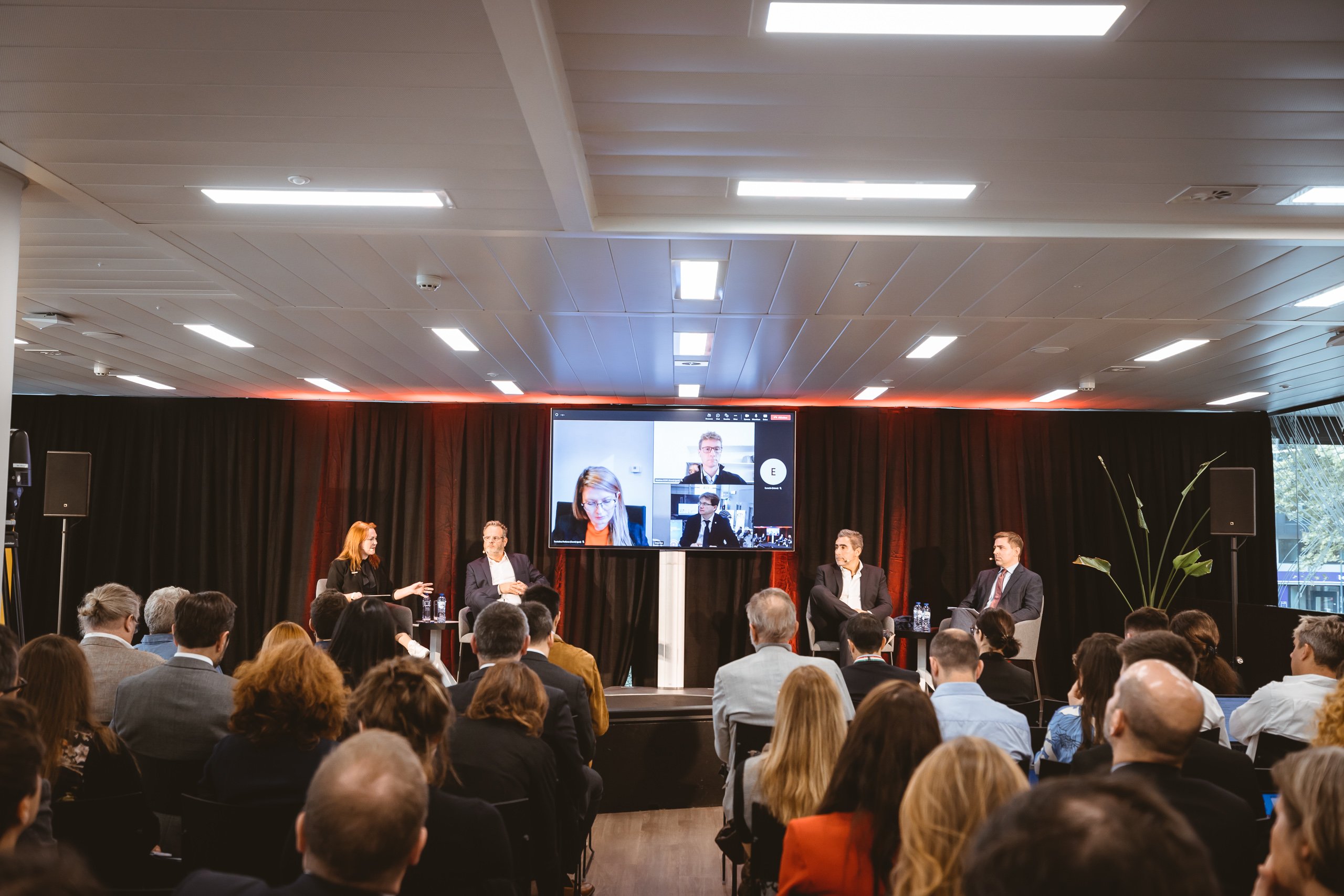 A conference panel with five speakers—three men and two women—sits onstage facing an audience. A large screen behind them displays remote participants in a video call. The room has modern lighting and a black curtain backdrop.