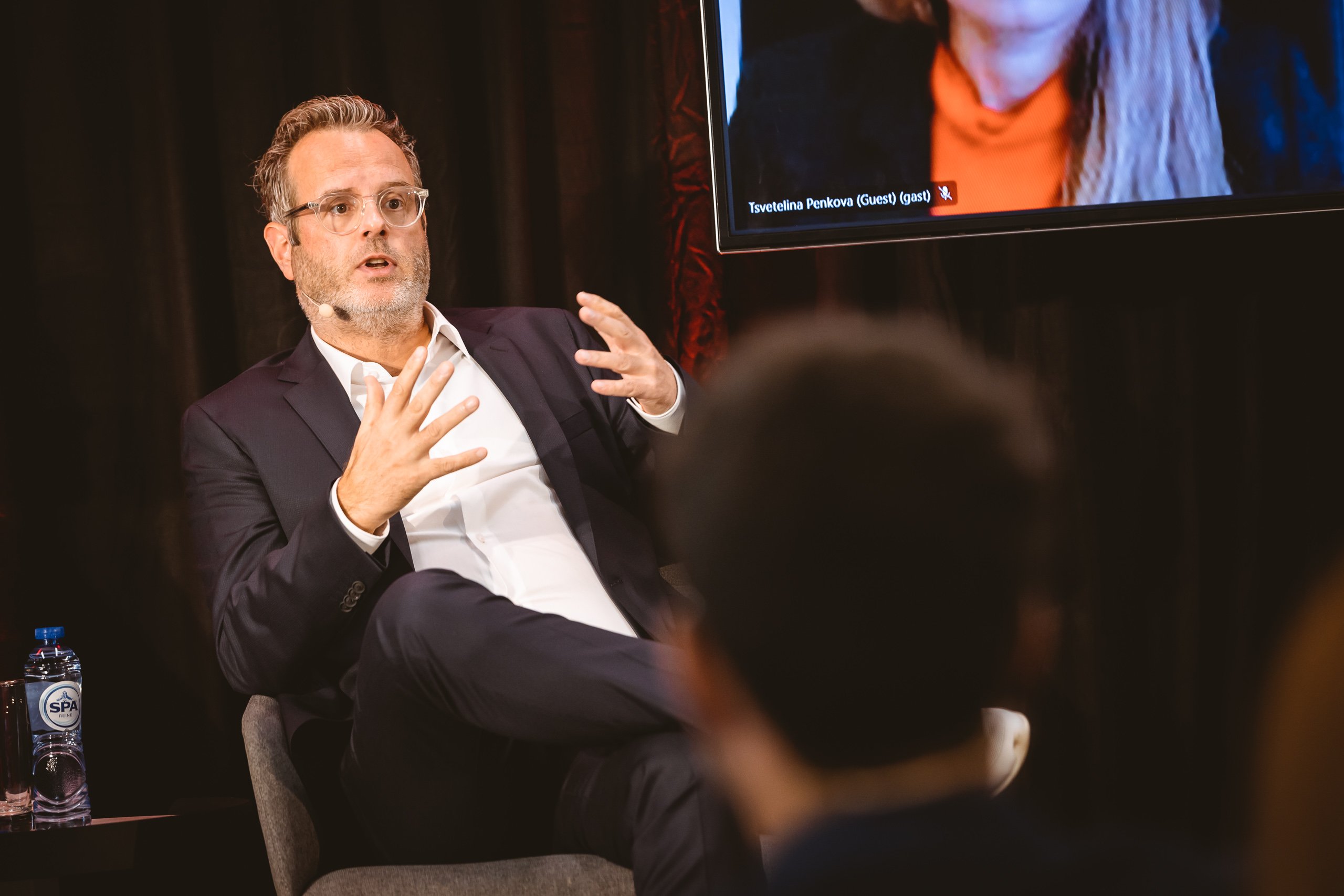 A man in a dark suit and glasses sits on a stage chair, gesturing with his hands while speaking. He wears a microphone headset. A bottle of water is placed next to him. In the background, part of a screen with a video call participant is visible.