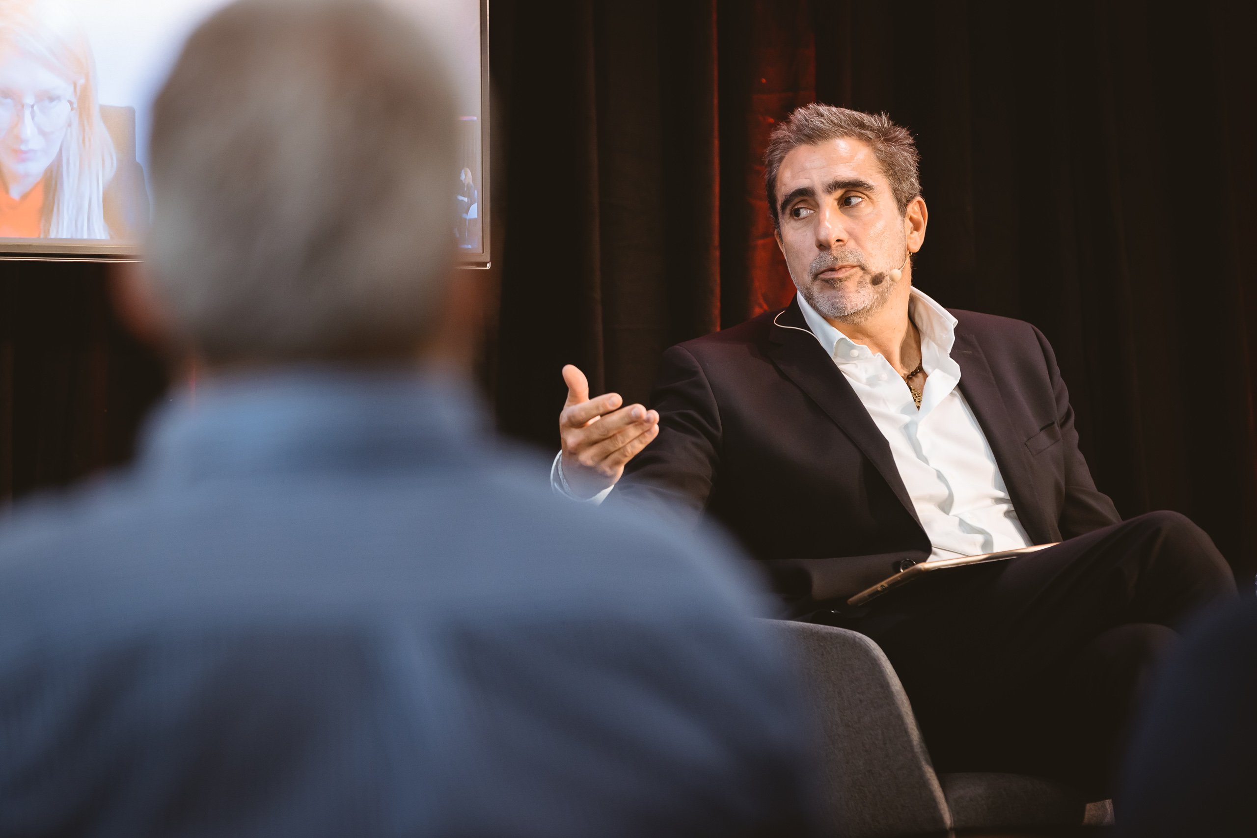 A man in a dark suit, sitting in a chair with a notepad, gestures while speaking on stage. He wears a microphone headset. The blurred figure of another person is in the foreground. A screen with a woman’s face is visible in the background.