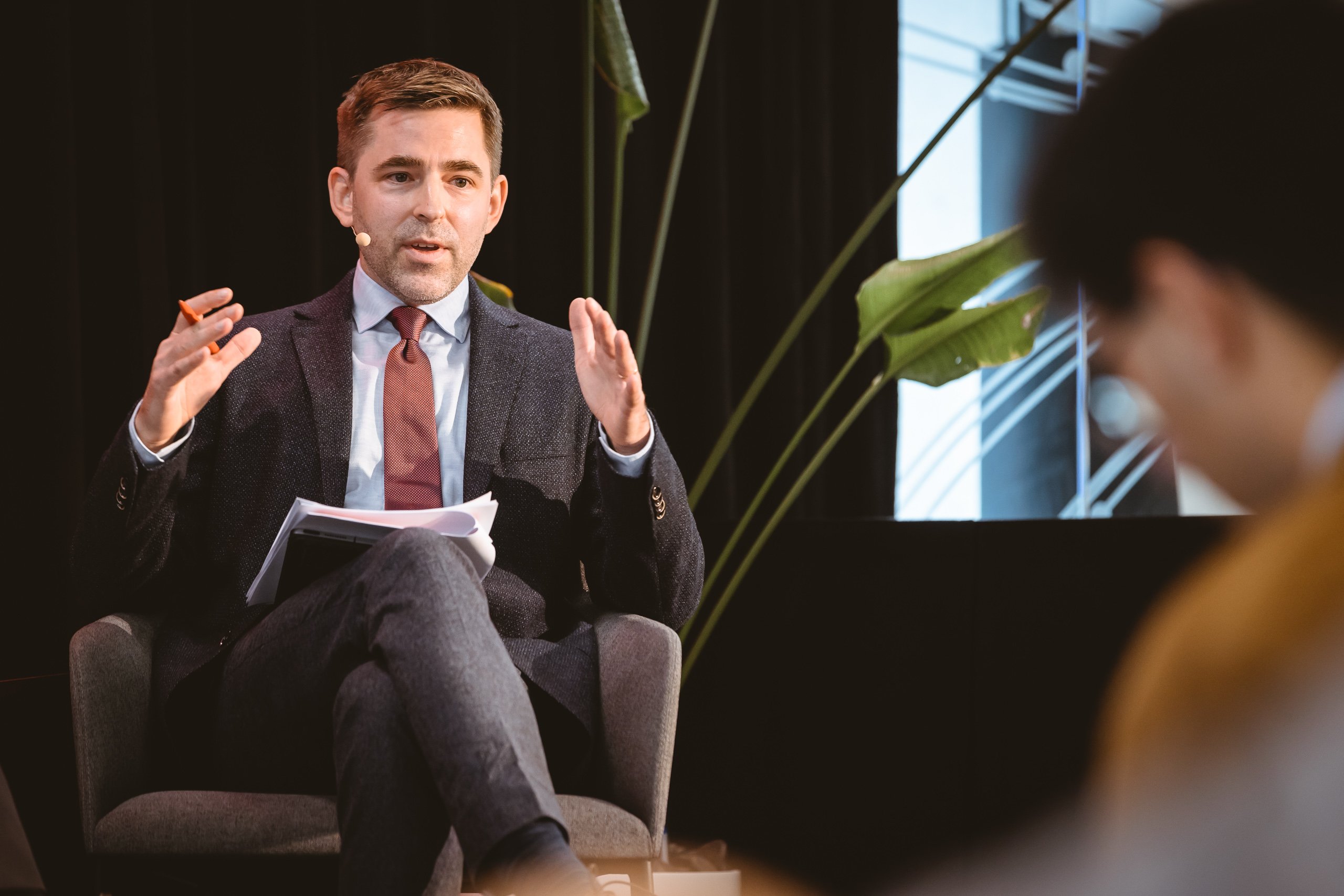 A man in a dark suit and red tie sits on a chair with legs crossed, gesturing as he speaks. He holds papers and wears a headset microphone. Large green plants and part of a modern building are visible in the background. Another person is blurred in the foreground.