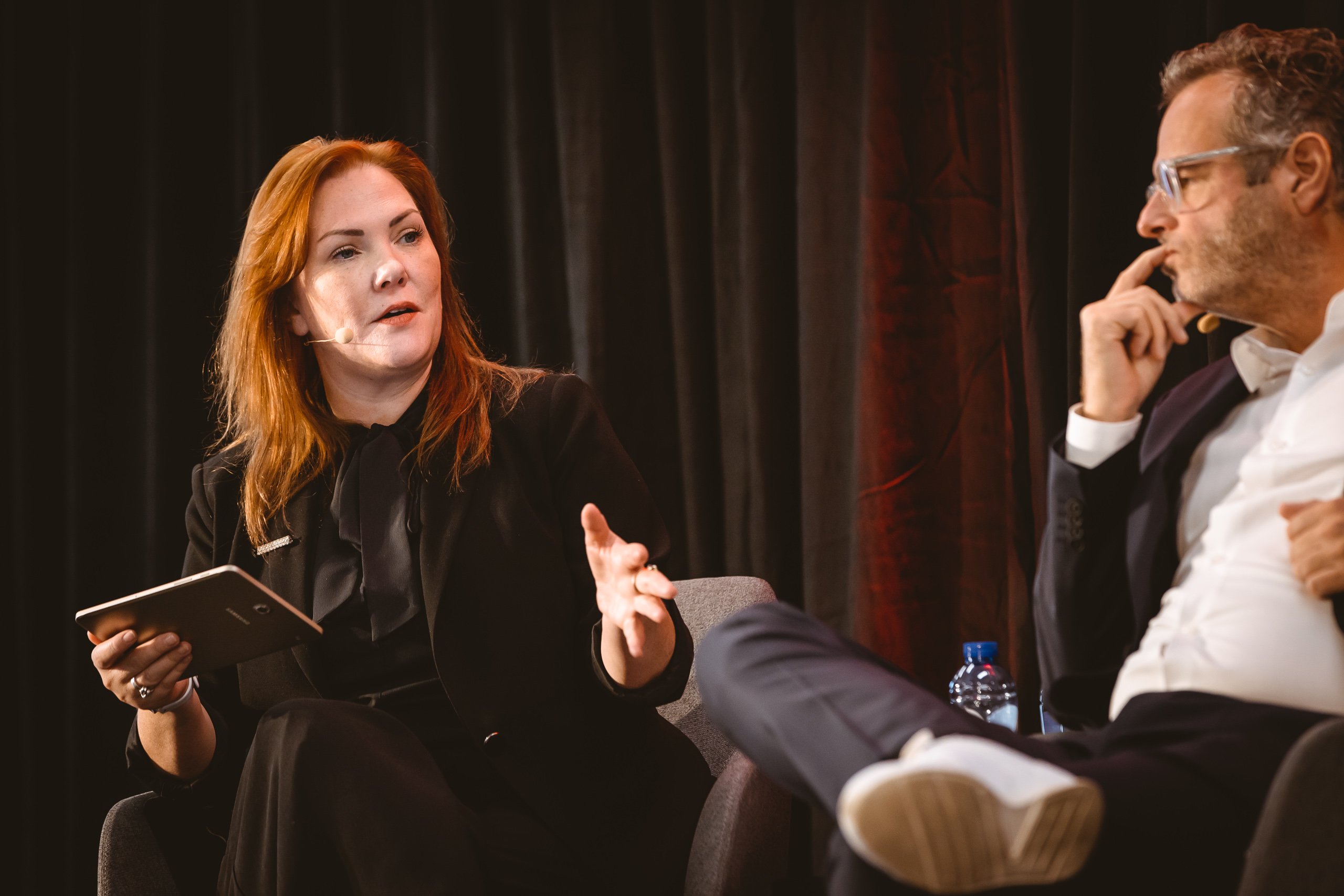 A woman with red hair speaks while holding a tablet, gesturing with her left hand. She wears a black suit and sits on a chair next to a man in glasses and a suit, who listens thoughtfully with his hand near his mouth. Both are onstage in front of dark curtains.