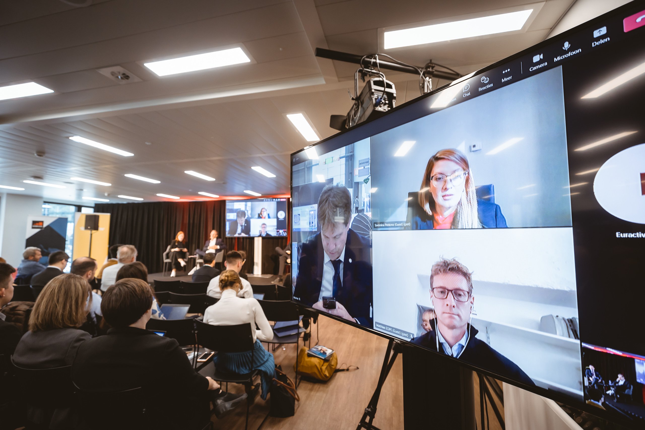 A conference room with seated attendees faces a stage where speakers sit. A large screen displays a video call with four participants, including two women and two men, appearing above the audience. The setting is modern and well-lit.
