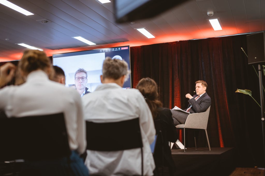 A speaker in a suit sits on stage in a gray chair, addressing an audience. Behind him, a large screen displays a man participating remotely via video call. Attendees are seated facing the stage, and the setting has modern lighting and dark curtains.