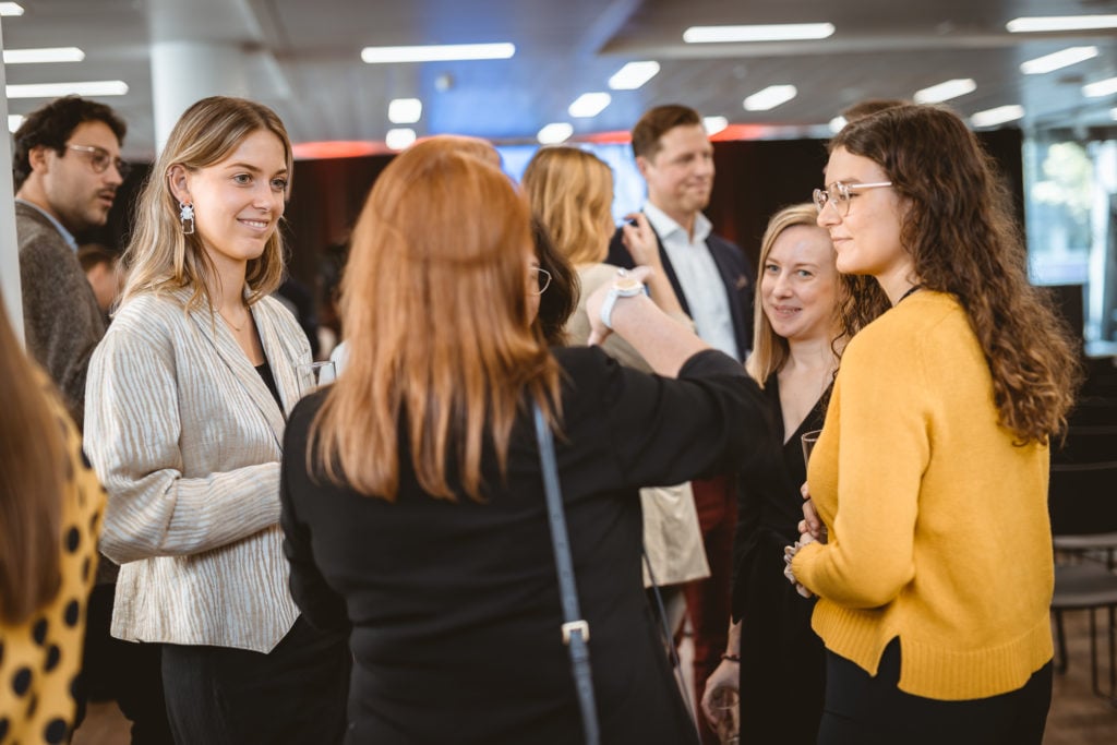 A group of people, mostly women, stand indoors in conversation. They are dressed in business casual attire, wearing blazers, sweaters, and glasses. The background is blurred, suggesting an event or conference setting with chairs and modern lighting.