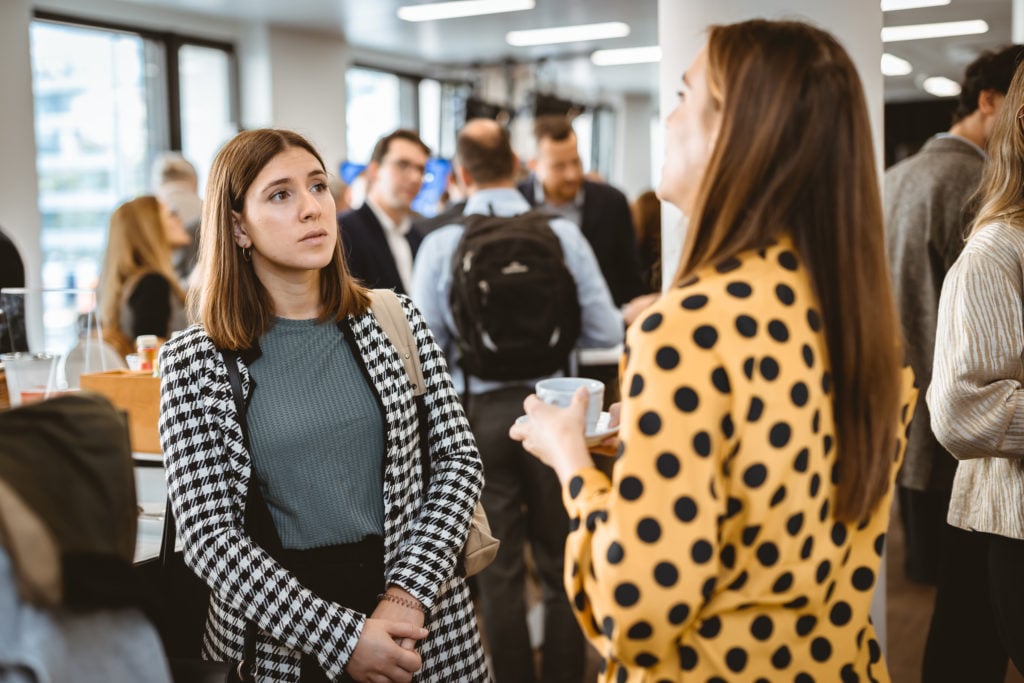 Two women converse at a networking event indoors. One wears a black-and-white houndstooth coat and listens attentively; the other wears a yellow polka-dot shirt and holds a cup. Several people mingle and chat in the blurred background.