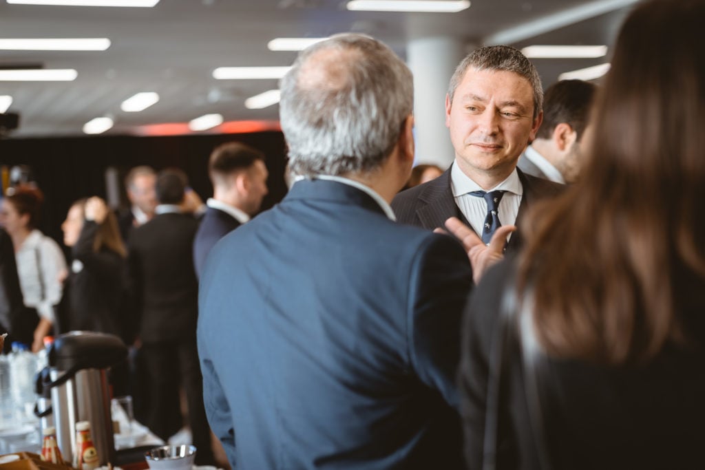 A group of people in formal attire converse at a networking event indoors. The focus is on two men talking; one faces the camera, wearing a suit and striped tie, while the other has his back turned. Blurred attendees and refreshments are visible in the background.