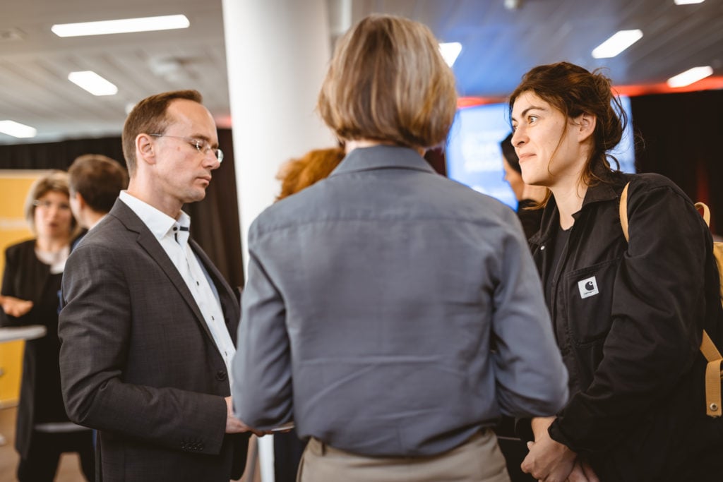 Three people stand in conversation at an indoor event. A man in a suit speaks to two women, one with short hair seen from behind, and another with dark hair and a black jacket. In the background, other attendees and large screens are visible.