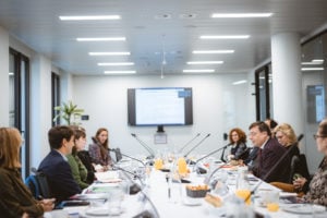 A group of people sits around a long conference table in a modern, bright meeting room. Papers, drinks, and snacks are on the table. A wall-mounted screen displaying text is visible in the background. The atmosphere appears focused and professional.