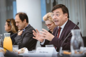 A man in a suit and glasses speaks passionately into a microphone at a conference table, gesturing with his hands. Other attendees sit beside him, listening. The table has name tags, glass pitchers of water and juice, and some food. The background is office-like.