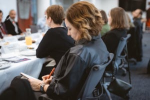 A woman with wavy, shoulder-length hair sits at a conference table, writing in a notebook with a red pen. Several other people sit in a row beside her, engaged in discussion. The setting is formal, with white tablecloths and documents on the table.