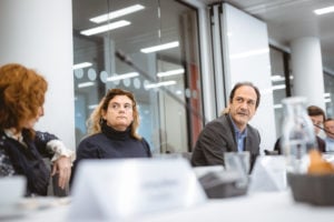 Three people sit at a conference table in a modern office with glass walls. The woman in the middle with curly hair looks attentive, while the man next to her speaks. White nameplates, glassware, and a pitcher are visible in the foreground.