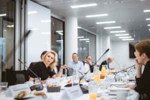 A group of people sit around a modern conference table with microphones, nameplates, drinks, and breakfast foods. One woman in black is speaking, while others listen attentively. The room is bright with glass walls and white lighting.