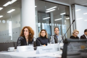 Four people sit at a conference table with nameplates, microphones, and water carafes. A woman with curly hair speaks while others listen. The room has glass walls, modern decor, and bright overhead lighting, suggesting a formal business meeting.