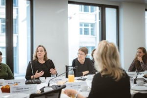 Five people are seated around a conference table in a modern, bright room with large windows. One woman speaks while gesturing with her hands. Name cards, coffee cups, and a pitcher of orange juice are on the table. Others listen attentively.