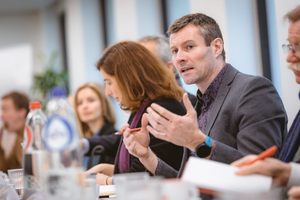 A man in a gray blazer speaks and gestures with his hands during a meeting at a conference table. Several people sit beside him, listening and taking notes. Bottles, papers, and pens are visible on the table in a bright office setting.