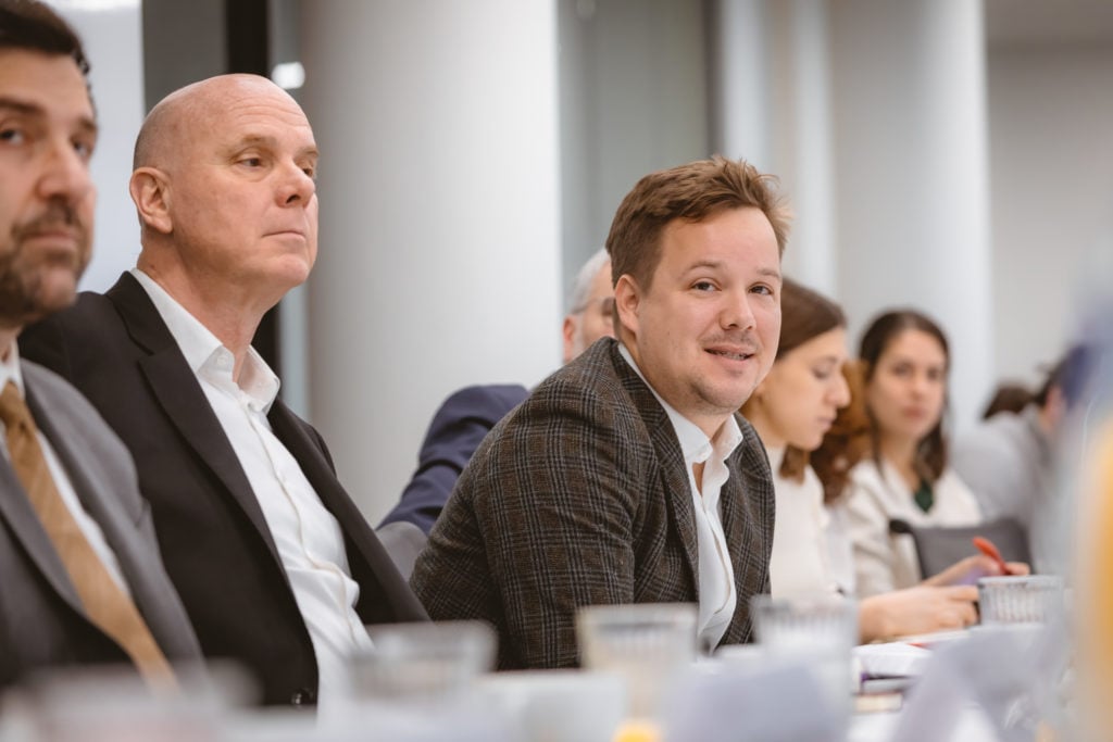 A group of professionals sits at a conference table in a meeting room. The focus is on a man in a plaid jacket who is smiling slightly, while others nearby listen attentively. Papers and cups are visible on the table in the foreground.