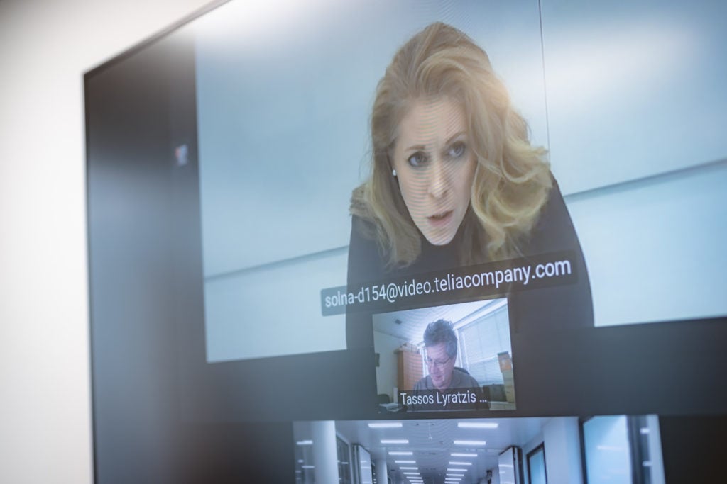 A woman with long blonde hair appears concerned during a video call, displayed on a large screen. Below her, a smaller window shows a man with glasses. Their emails and names are visible. A hallway with ceiling lights is reflected on the screen.