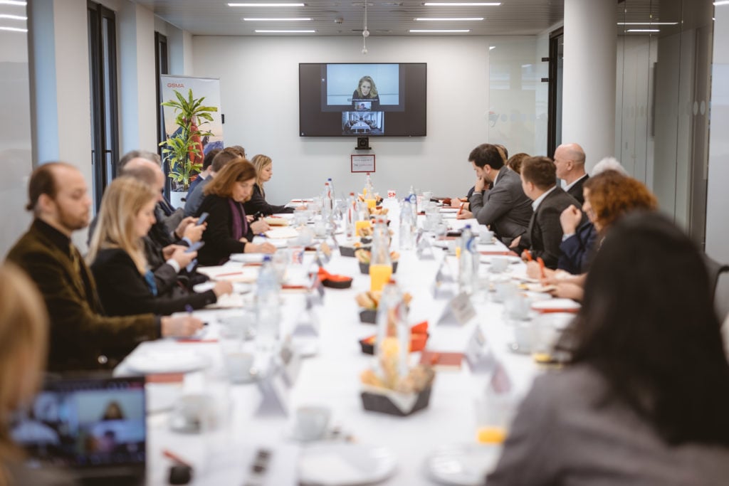 A group of people sit around a long conference table set with papers, drinks, and snacks. Most attendees are focused on a video call projected at the end of the room, where a woman appears on a large screen, leading the discussion.