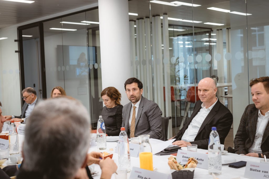 A group of people in business attire sits around a conference table with documents, nameplates, drinks, and snacks. They are engaged in discussion in a modern glass-walled office with natural light.