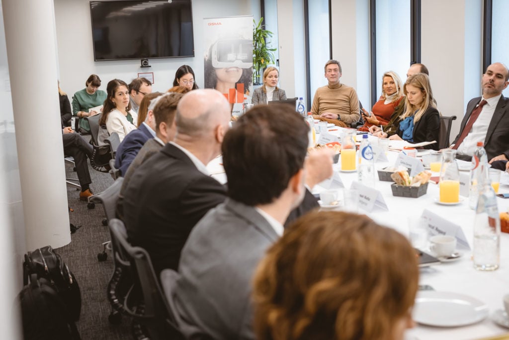 A group of people sit around a long conference table set for a meeting, with orange juice, pastries, and name cards visible. Some participants are engaged in conversation, while others listen. Large windows and a TV screen are in the background.