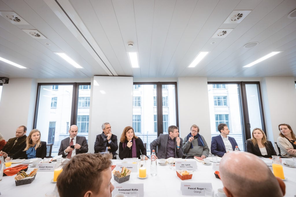 A group of people in business attire sit at a long conference table with nameplates, drinks, and breakfast items in front of large windows. They are engaged in conversation in a modern, well-lit meeting room.