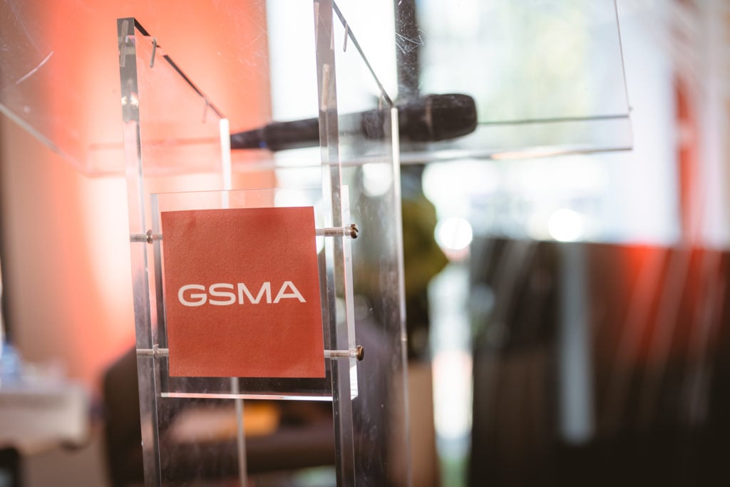 A clear acrylic podium with a red sign displaying the white letters "GSMA" is shown. The background is softly blurred, with warm light and faint shapes of windows and possibly greenery outside.