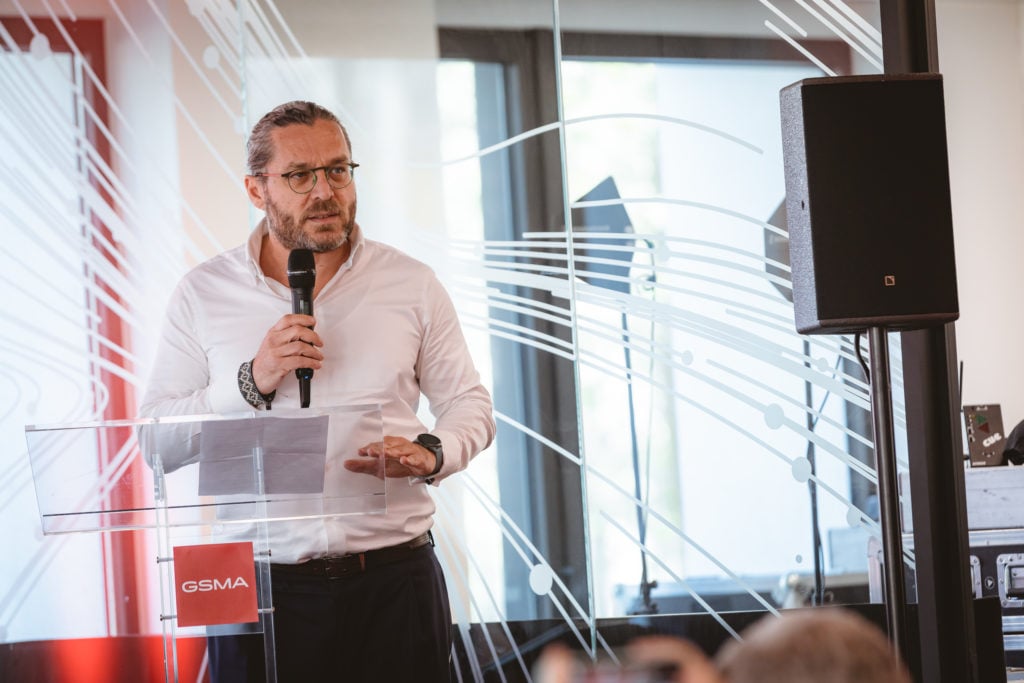 A man with glasses and a white shirt speaks into a microphone at a podium labeled "GSMA." He stands in a modern room with large windows, abstract white line patterns on glass, and a black speaker visible beside him.