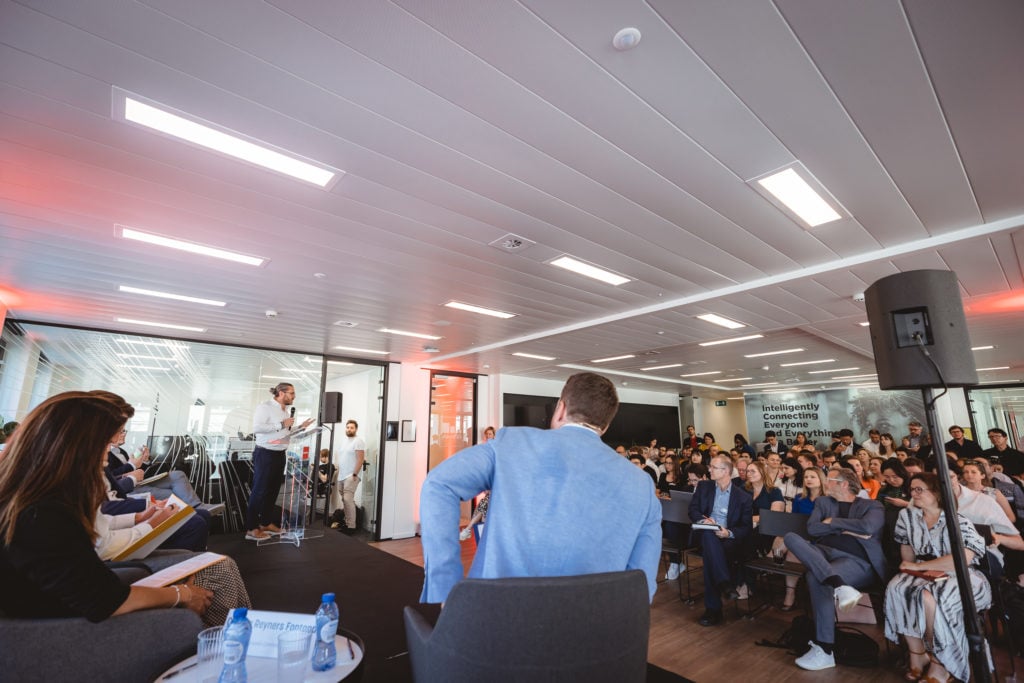 A man stands at a podium speaking to a seated audience in a modern conference room. Attendees listen attentively; some take notes. A woman sits on stage with documents, and a man in a blue jacket faces the crowd. A sign on the back wall reads “Reimagining Cement.”.