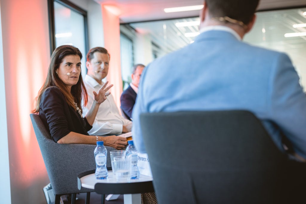 A woman with long brown hair sits in a gray chair, gesturing as she speaks during a panel discussion. Three men sit beside her, and another man in a blue suit faces away in the foreground. Water bottles are on the table; soft, warm lighting fills the modern room.
