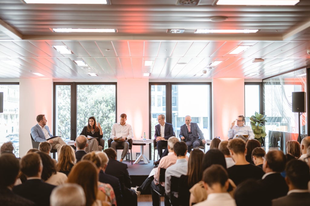 A panel of six people, four men and two women, sit on a stage in front of large windows, speaking at a professional event. The audience faces them, and the room is brightly lit with modern decor and ceiling lights.