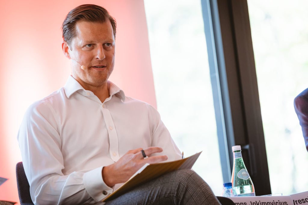 A man with short, slicked-back hair, wearing a white dress shirt and patterned pants, sits indoors holding a folder. He has a headset microphone and gestures with his hand. A bottle of water and a blurred nameplate are on a table beside him.